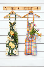 Two colorful aprons hanging on wooden hangers from a wooden peg rack against a white wood-plank wall. The left apron has a green, yellow, and white floral print with a pocket holding carrots and greens.
