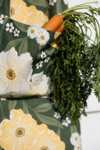 A close-up of a dark green apron with a large floral print of white daisies and yellow flowers, featuring a side pocket holding a fresh carrot with green tops.