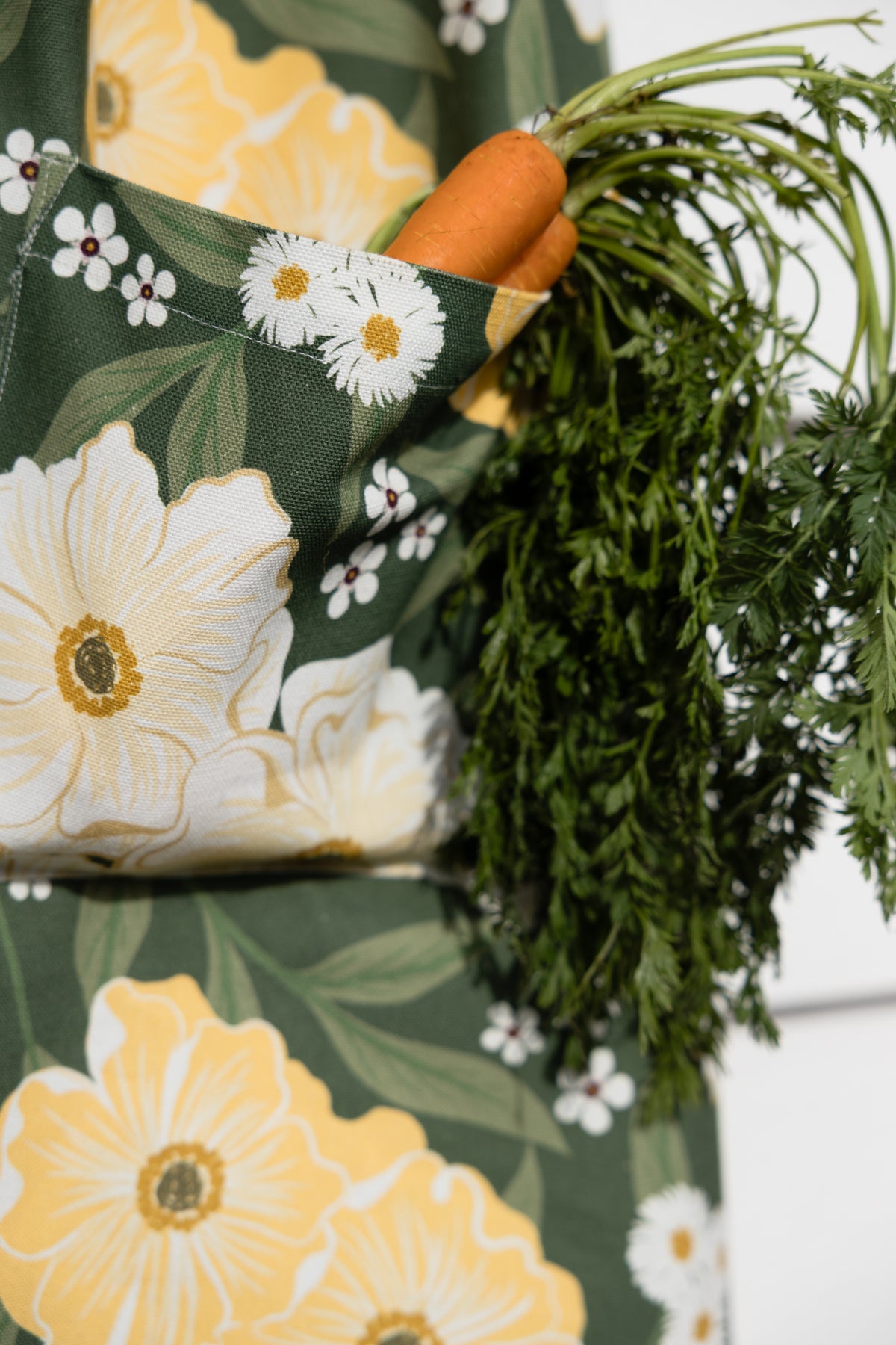 A close-up of a dark green apron with a large floral print of white daisies and yellow flowers, featuring a side pocket holding a fresh carrot with green tops.